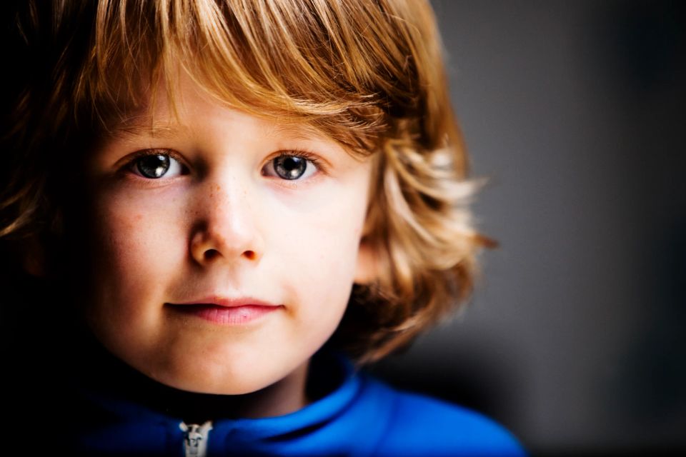 Nærbillede af et barn med lyst hår og udtryksfulde øjne, iført en blå bluse, i blødt sidelys. Portrætfoto.