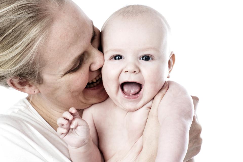 Glad baby griner højt i fokus, holdt af en kvinde med lyst hår, der smiler ømt. Et babyfotografi med hvid baggrund.