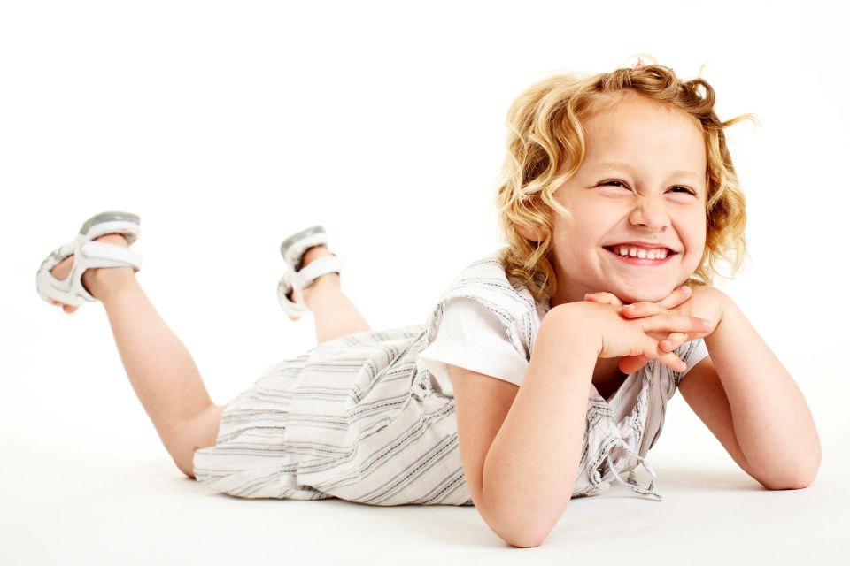 Børnefotografering af glad lyshåret barn der ligger på maven, smilende og kigger mod kameraet i legende stemning.