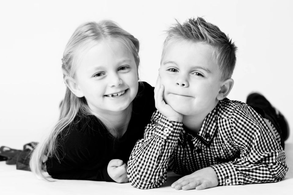 Sort/hvid studieportrætfoto af to smilende børn, en pige og en dreng, der ligger tæt sammen og kigger i kameraet.