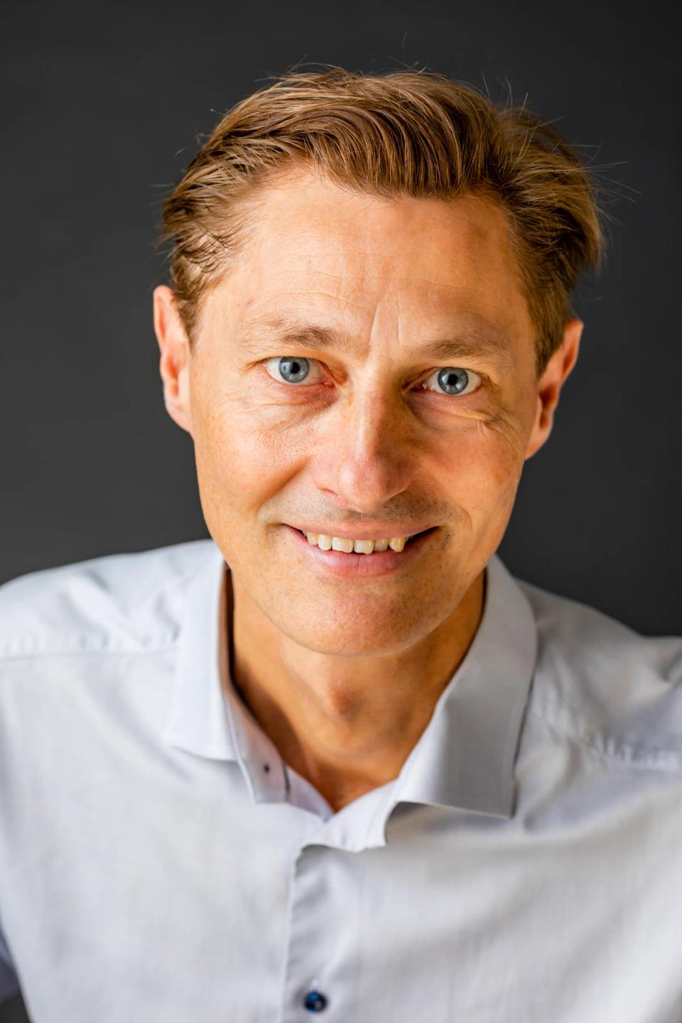 Nærbillede af smilende mand i hvid skjorte med blå øjne og lyst hår, et professionelt portrætfoto med blødt lys.
