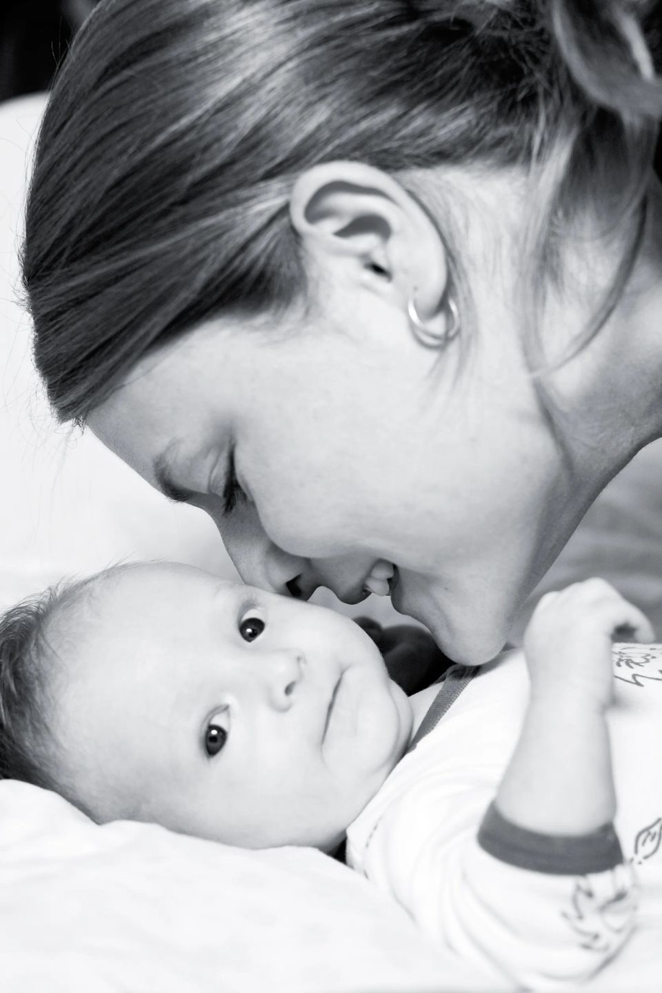 Sort/hvid babyfoto af mor der kysser sit barn på panden med rørende nærhed. Babyen kigger nysgerrigt frem.