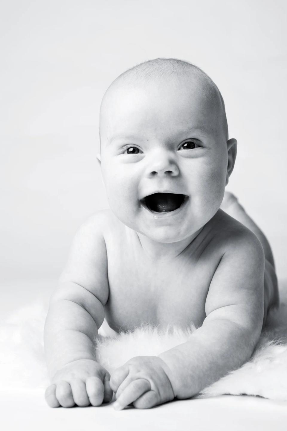 Sort-hvid babyfotografering: Glad baby griner liggende på et hvidt, blødt tæppe med åben mund og strålende øjne.
