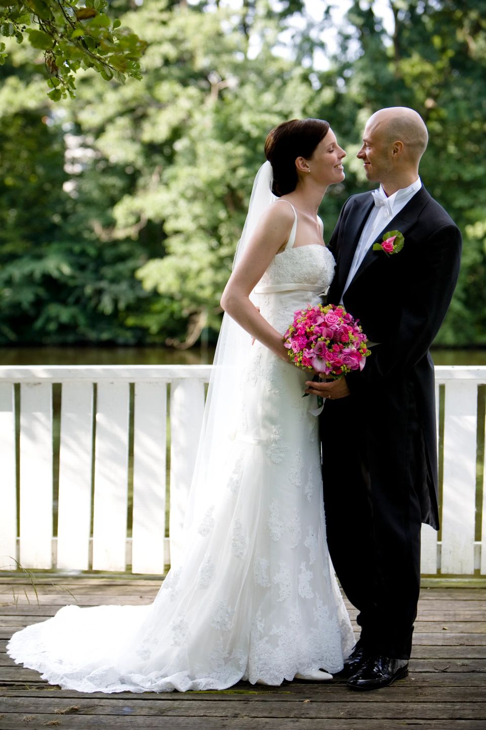 Bryllupsfotografi af smilende brudepar der ser hinanden dybt i øjnene, stående på træterrasse ved vandet.