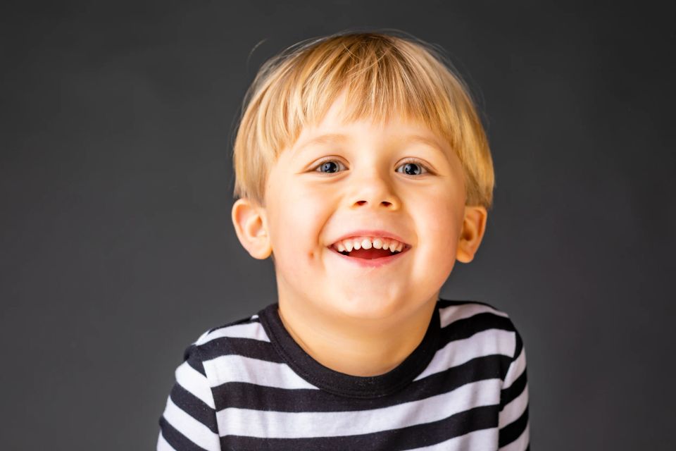 Nærbillede af smilende, glad dreng med lyst hår i sort-hvid stribet trøje mod mørk baggrund. Et livligt børneportræt.