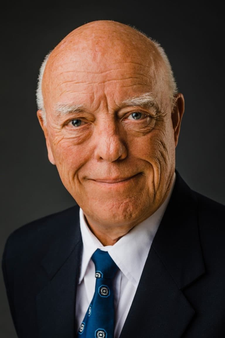 Nærbillede af ældre, smilende mand i sort jakkesæt og blåt slips, der poserer for et autentisk portrætfoto.