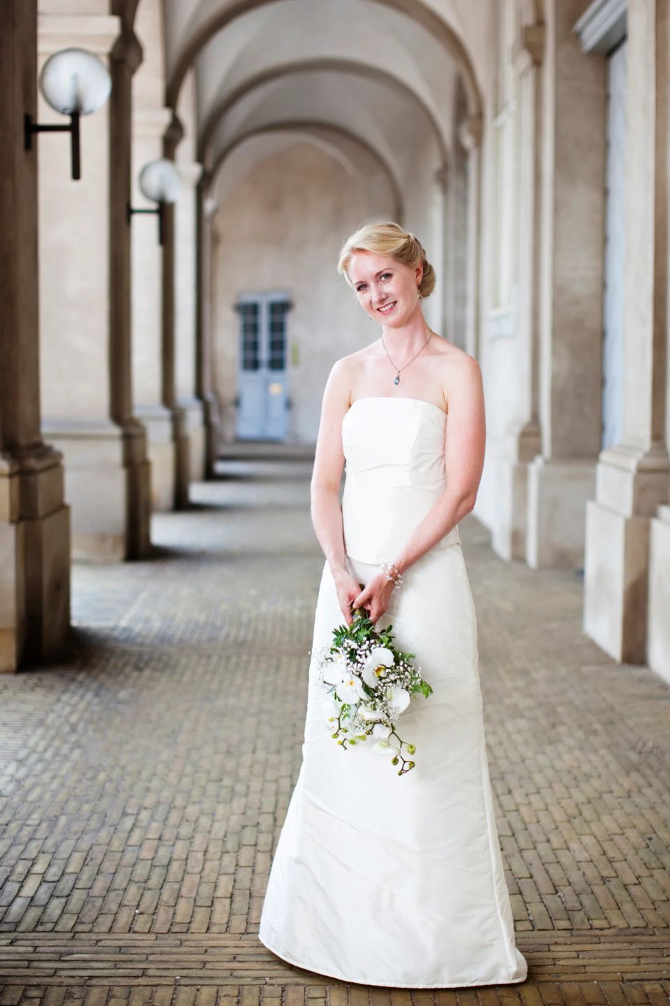 Bryllupsfotografering af smilende brud i hvid kjole med brudebuket på Christiansborg Ridebane. Lyse, glade farver.