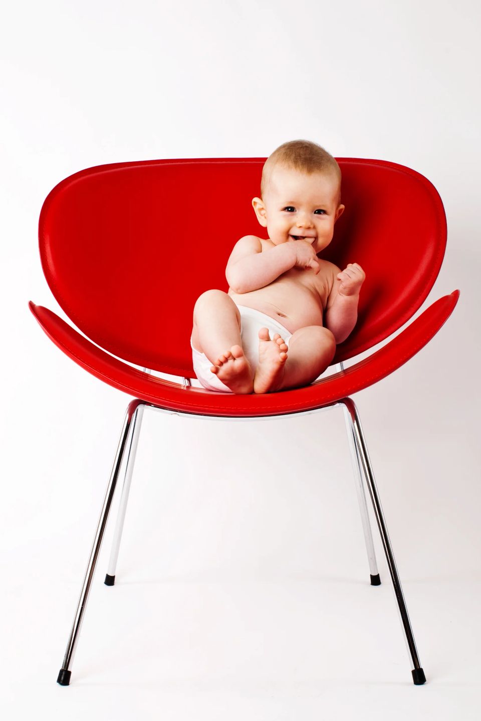 Glad baby i hvid ble smiler og sidder i en rød stol mod hvid baggrund. Dejligt babyfotografi med naturligt lys.