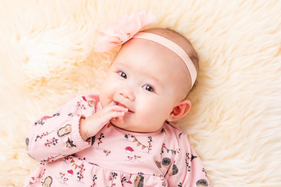 Nærbillede af smilende baby i en lyserød kjole med et blomstret hårbånd, liggende på et lunt, lyst tæppe. Babyfotografering.