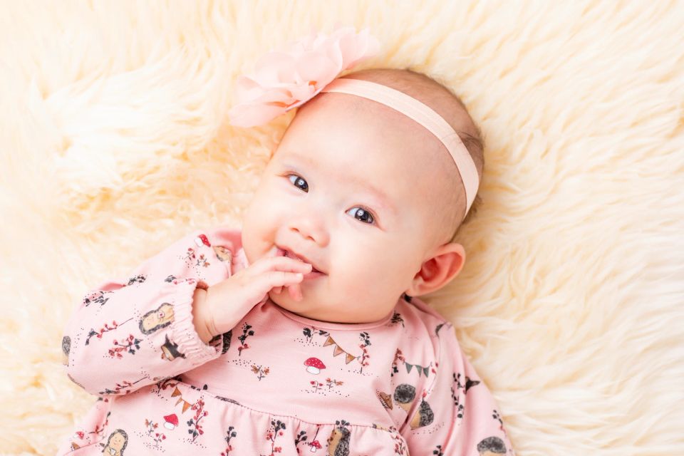 Nærbillede af smilende baby i en lyserød kjole med et blomstret hårbånd, liggende på et lunt, lyst tæppe. Babyfotografering.