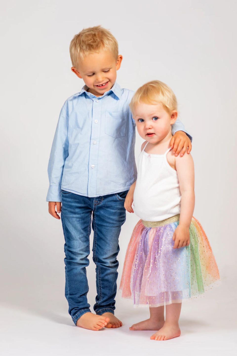 Legende børn: Stor dreng i lyseblå skjorte holder om lillesøster i farverig tylskørt, begge smiler, mod hvid baggrund.