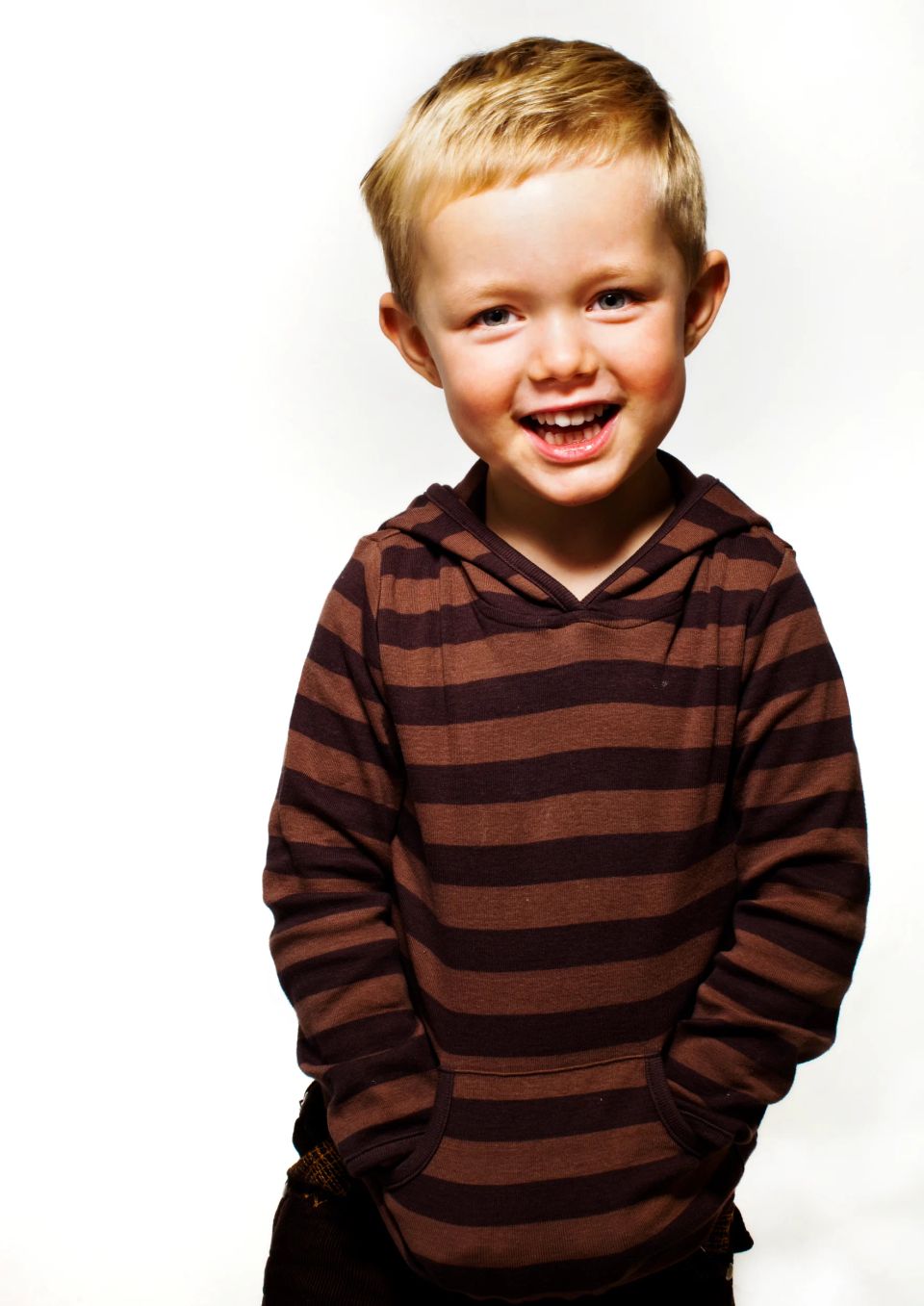 Glad, smilende dreng i en brun og mørkebrun stribet hættetrøje med hænderne i lommen, fotograferet mod hvid baggrund i blø...