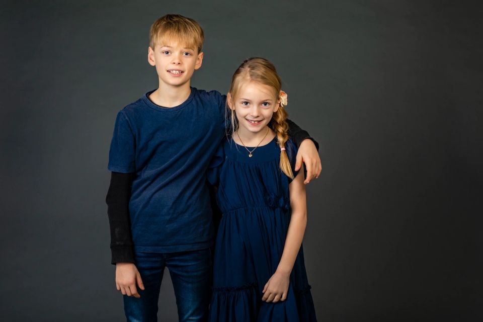 Familiefoto af smilende søskende, en dreng og en pige, der krammer mod en mørkegrå baggrund. Begge i mørkeblåt tøj.
