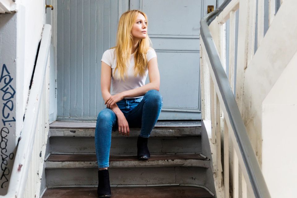 Portrætfoto af en ung blond kvinde i hvid T-shirt og jeans, der sidder på en gammel trætrappe og kigger op. Jeg fanger ste...