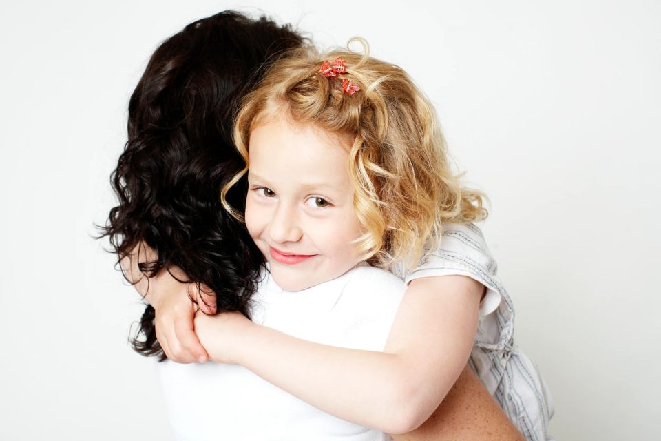 Familiefotografering: Smilende pige med lyst krøllet hår i kram med sin mor, taget tæt på i et blødt lys.