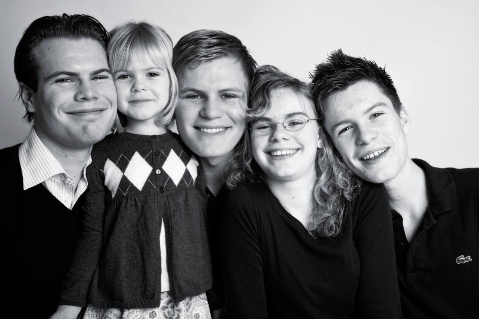 Autentisk familieportræt i sort-hvid med fem familiemedlemmer smilende mod kameraet i blødt lys.