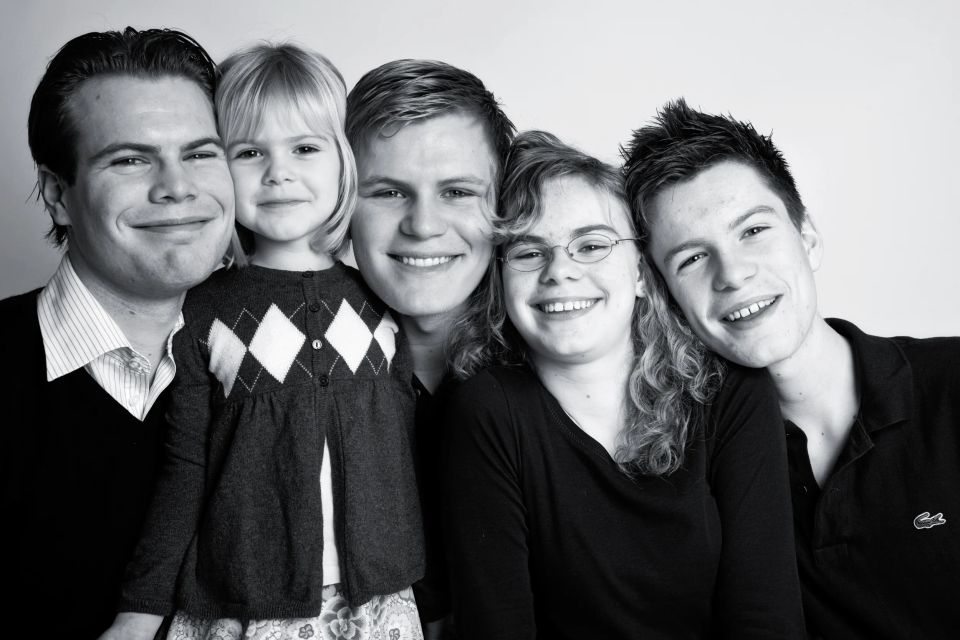 Autentisk familieportræt i sort-hvid med fem familiemedlemmer smilende mod kameraet i blødt lys.