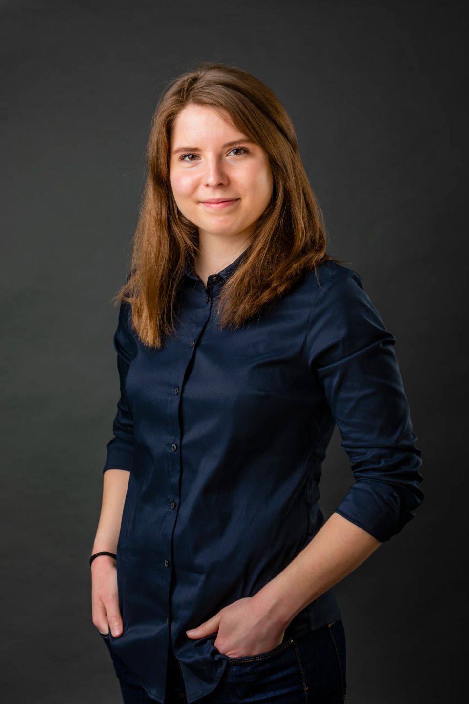 Portrætfoto af smilende kvinde med brunt hår i mørkeblå skjorte, stående i blødt lys mod mørk baggrund.