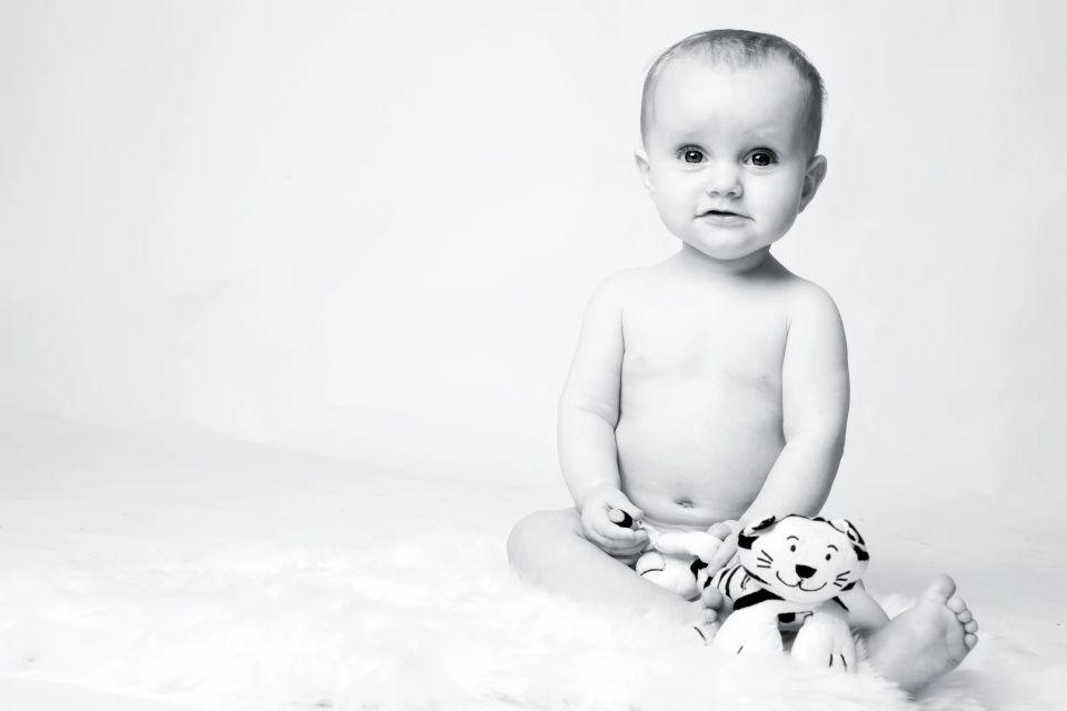 Babyfotografering af siddende baby mod hvid baggrund, kigger nysgerrigt med sit tøjdyr ved siden.
