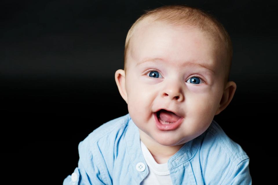 Nærbillede af glad baby med åben mund og strålende blå øjne i blåt tøj mod mørk baggrund. Et livligt babyfoto.