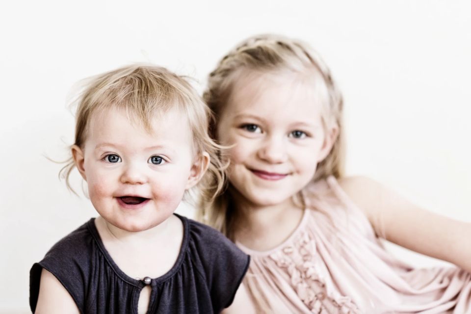 Børn: To smilende søstre fotograferet i naturligt lys. Den yngste kigger direkte mod kameraet med åben mund.