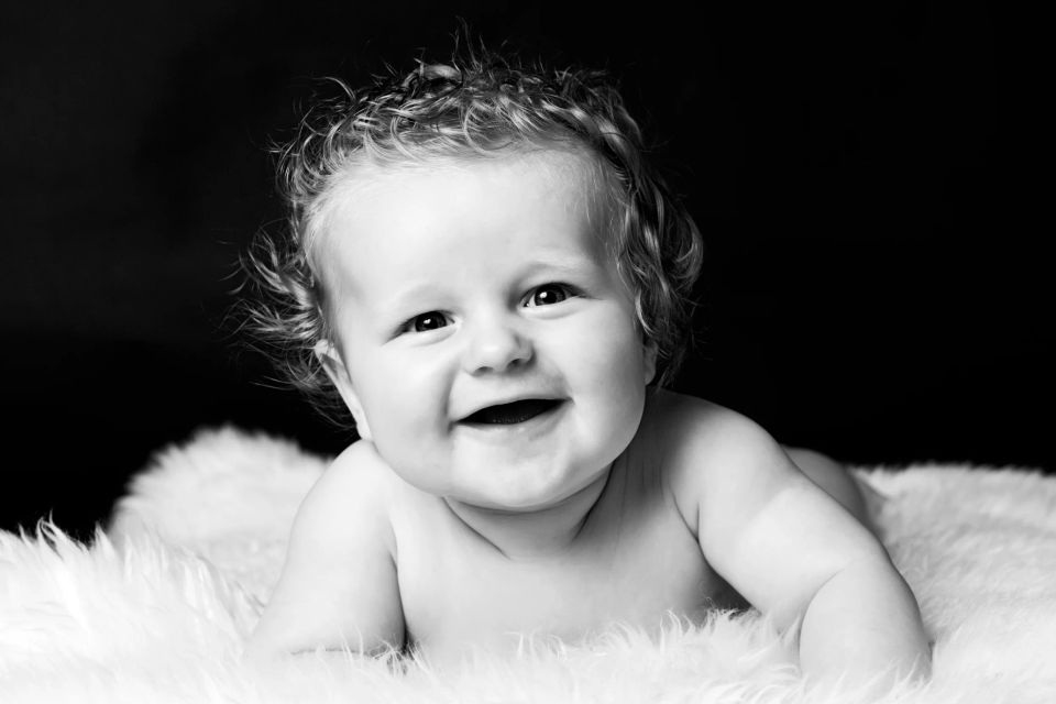 Babyportræt i sort-hvid af en smilende baby med krøllet hår, liggende på et hvidt, blødt tæppe. Foto i naturligt lys.