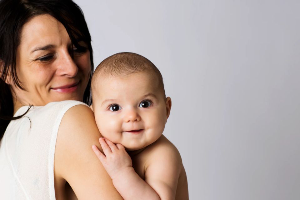 Glad baby fotografering. Baby kigger smilende frem over mors skulder i blødt studie lys.