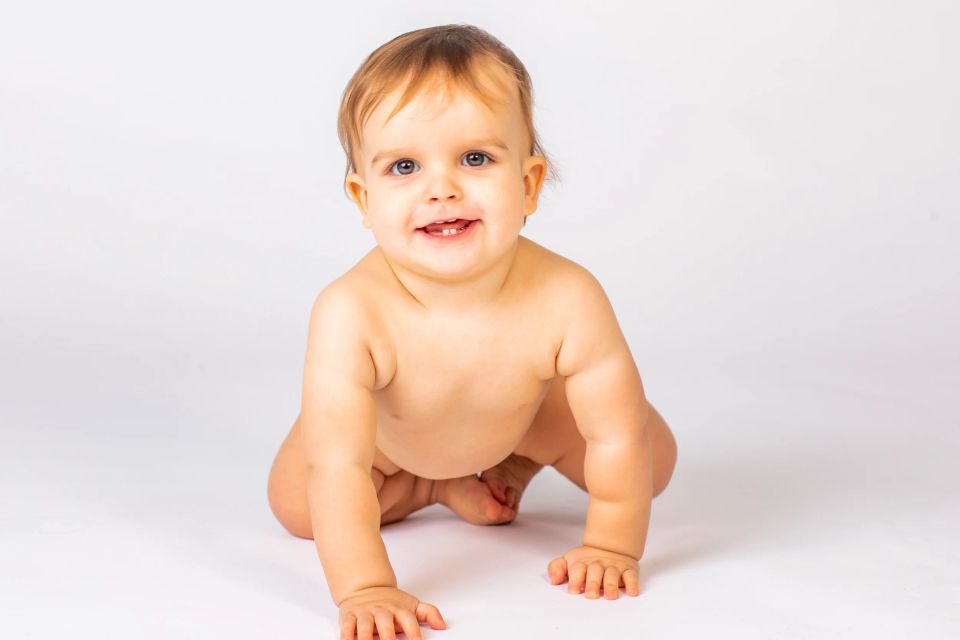 Glad baby kravler og smiler mod beskueren. Autentisk babyfotografering med hvid baggrund i naturligt lys.