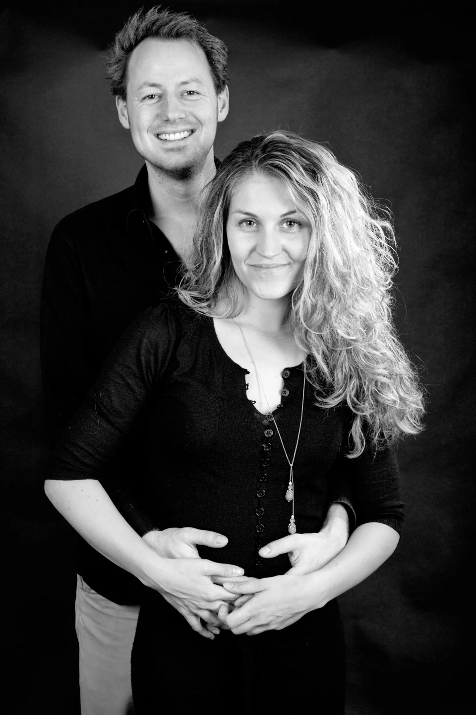 Familiefoto af smilende par, hvor manden står bag kvinden og holder hende om maven. Sorthvid graviditetsfotografering.