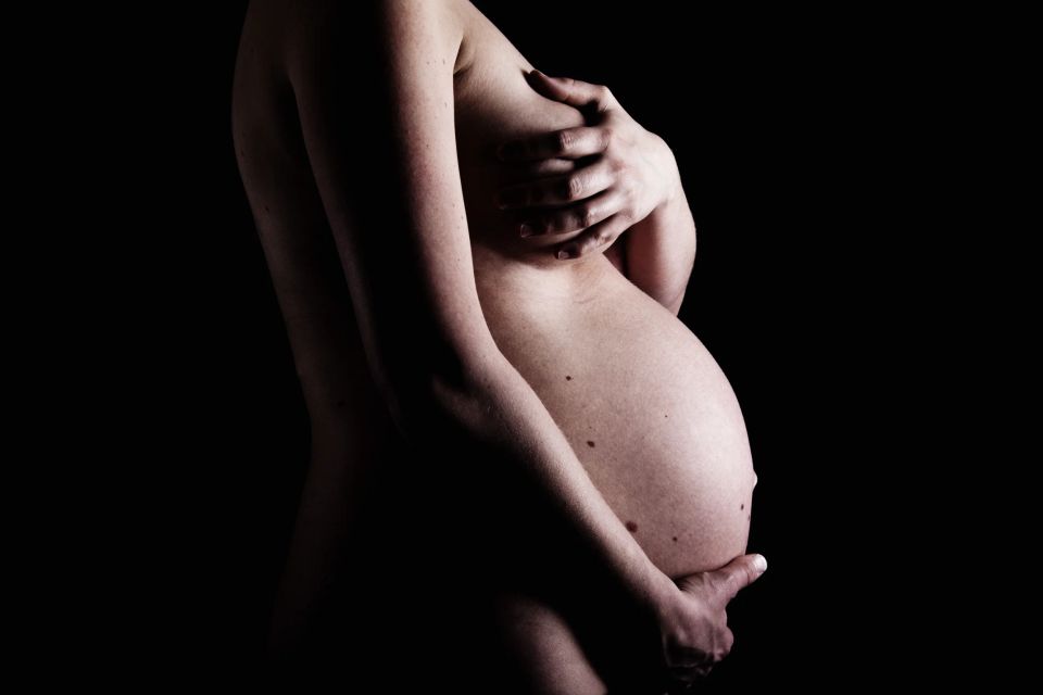 Gravid kvinde poserer sidelæns, holder kærligt om sin mave i blødt sidelys mod en mørk baggrund. Gravidfotografering.