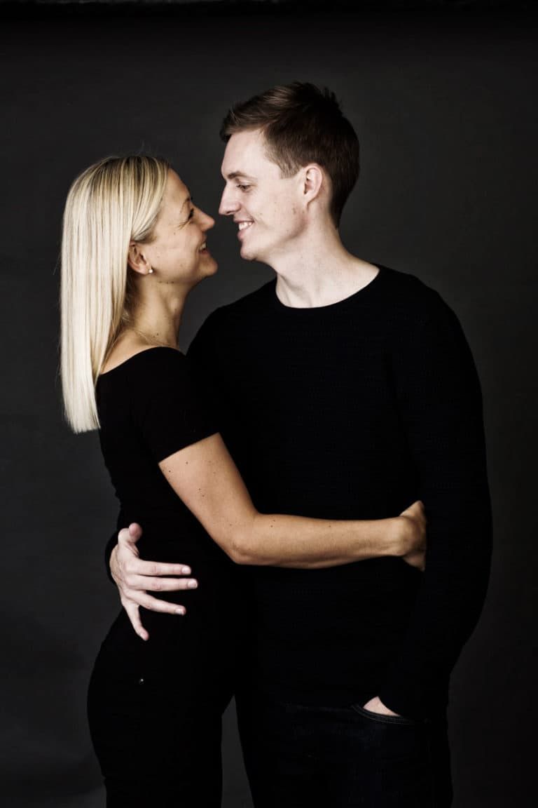 Portrætfoto af forelsket par der smiler og kigger kærligt på hinanden foran en mørk baggrund.