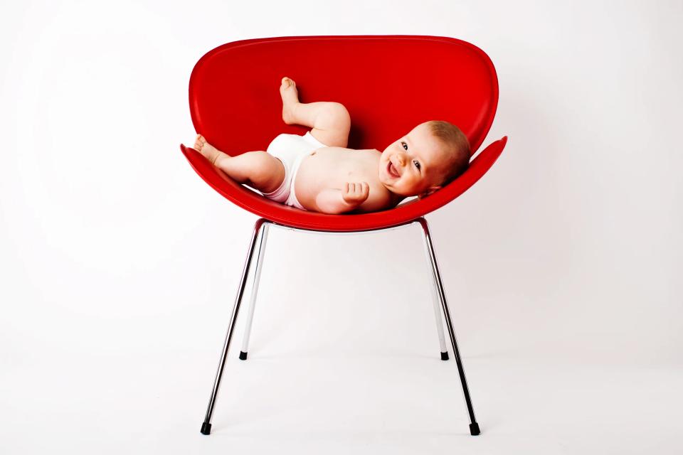 Glad baby i hvid ble liggende i en rød skalstol mod en lys baggrund. Følelse af glæde og leg iver.