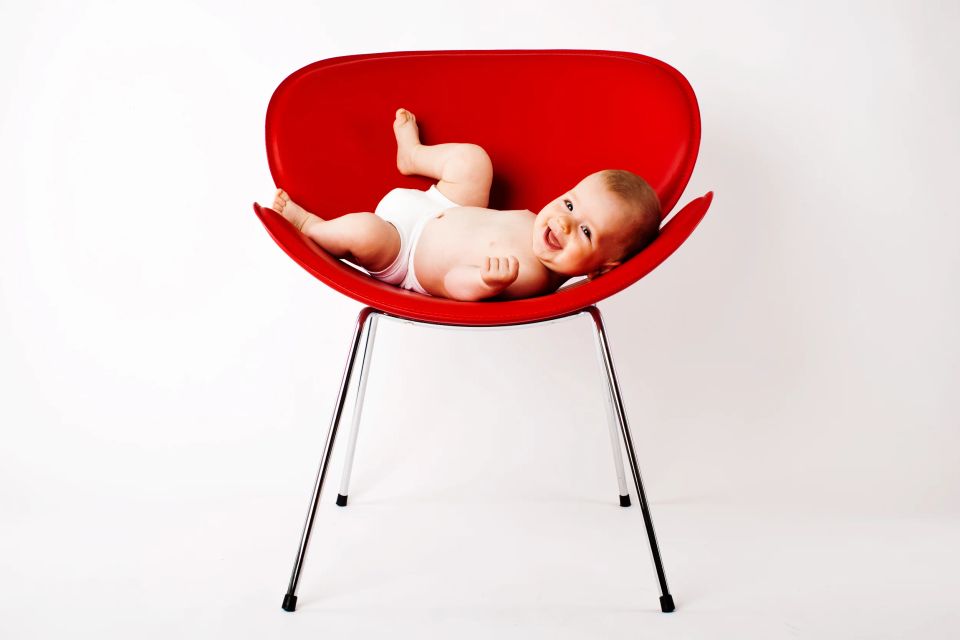Glad baby i hvid ble liggende i en rød skalstol mod en lys baggrund. Følelse af glæde og leg iver.