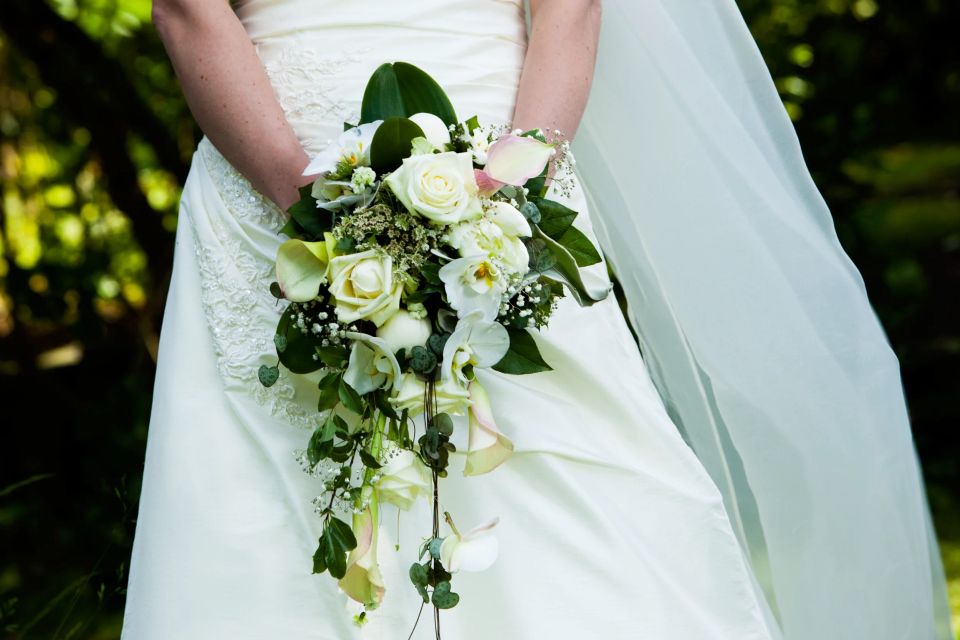 Nært bryllupsfoto af brudebuket med hvide roser og grønne blade holdt af bruden i hvid kjole og slør en lang lysegrøn slør.