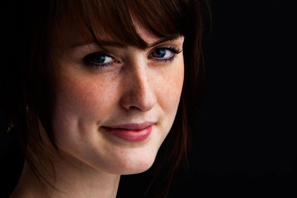 Nærbillede af ung kvinde med fregner, brunt hår og et varmt smil. Vinklen understreger personligheden i dette portrætfoto.