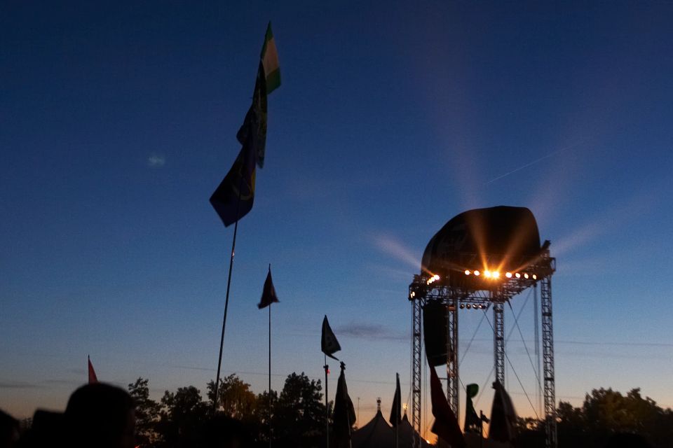 Aftenfoto af Roskilde Festival med scenelys der stråler mod den mørkeblå himmel, silhuetter af telte og flag i forgrunden.