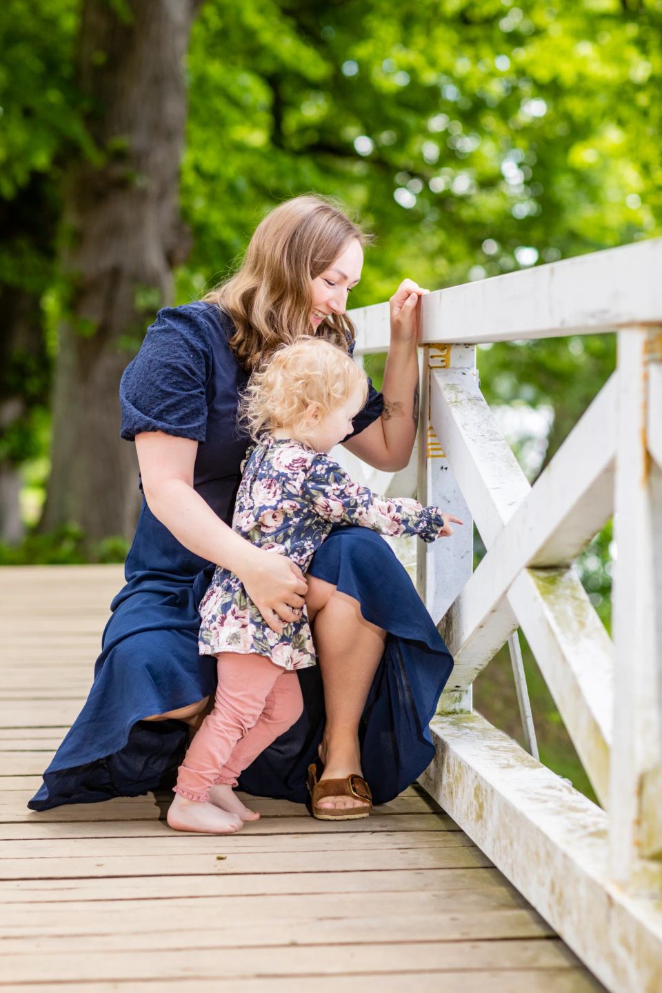 Mor og barn i blå kjoler på træbro. Barnet peger nysgerrigt ud mod grøn natur. Et roligt familieportræt.