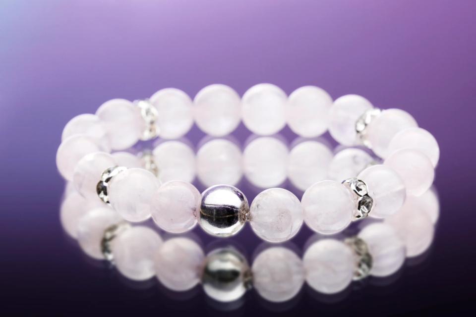 Elegant armbånd af lyserøde perler med en skinnende sølvperle og sølvdetaljer, reflekteret på en mørk baggrund. Et smukt a...