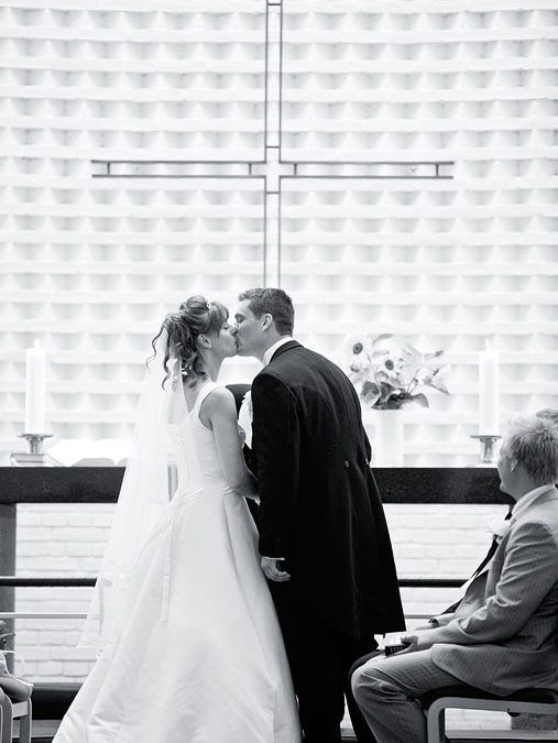 Bryllupsfotografering af brudepar, der kysser hinanden foran et kors i kirken i sort-hvid.