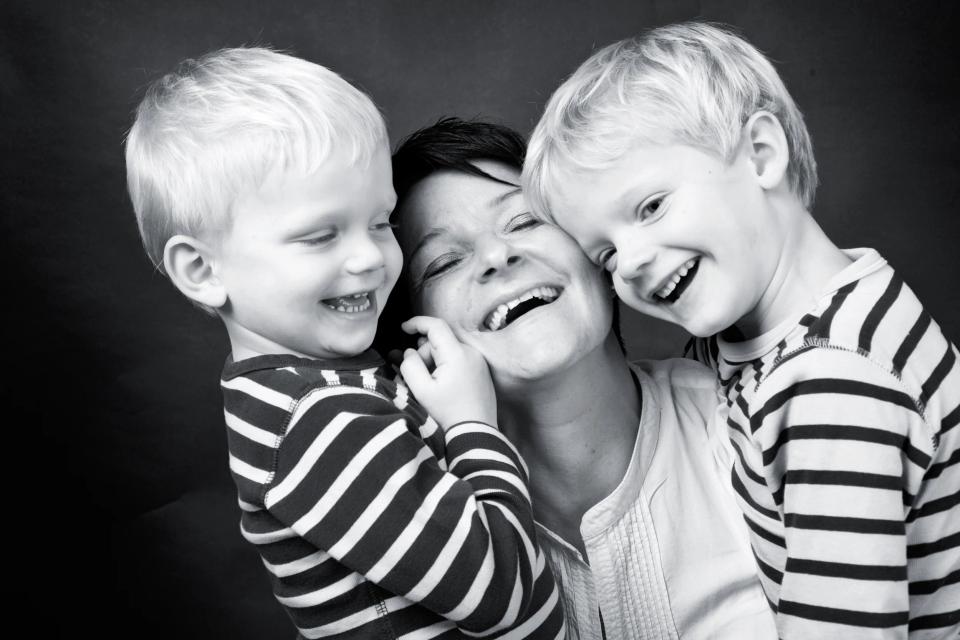 Sort/hvid familiefoto af mor og to små drenge, der griner og omfavner hinanden tæt med stor glæde.