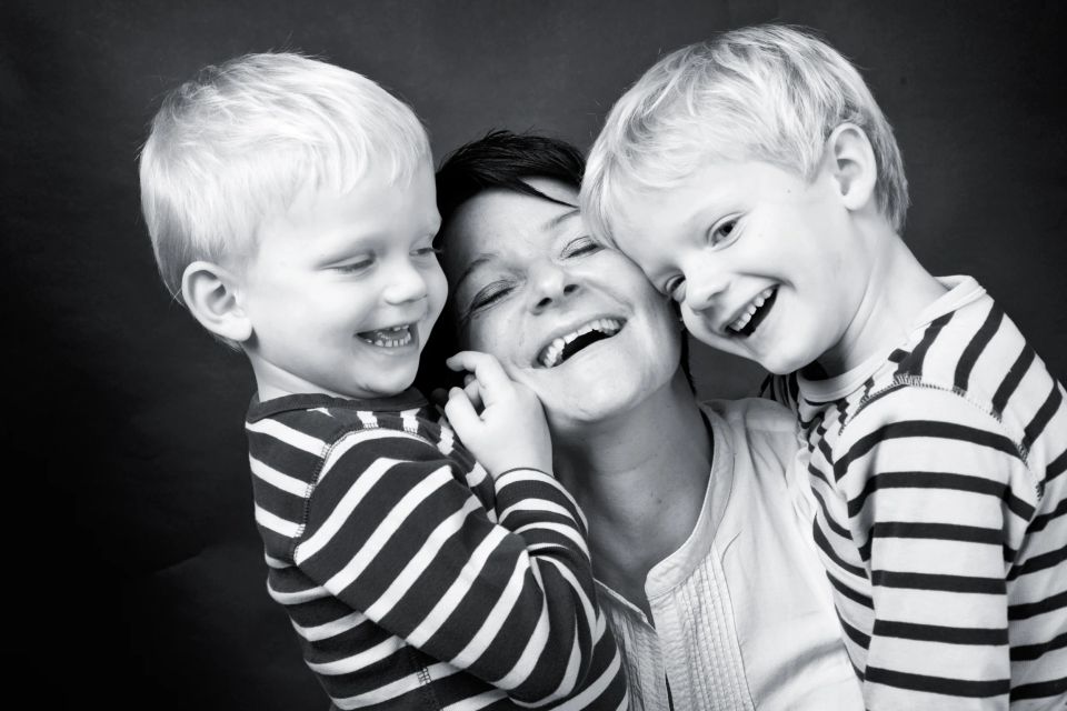 Sort/hvid familiefoto af mor og to små drenge, der griner og omfavner hinanden tæt med stor glæde.