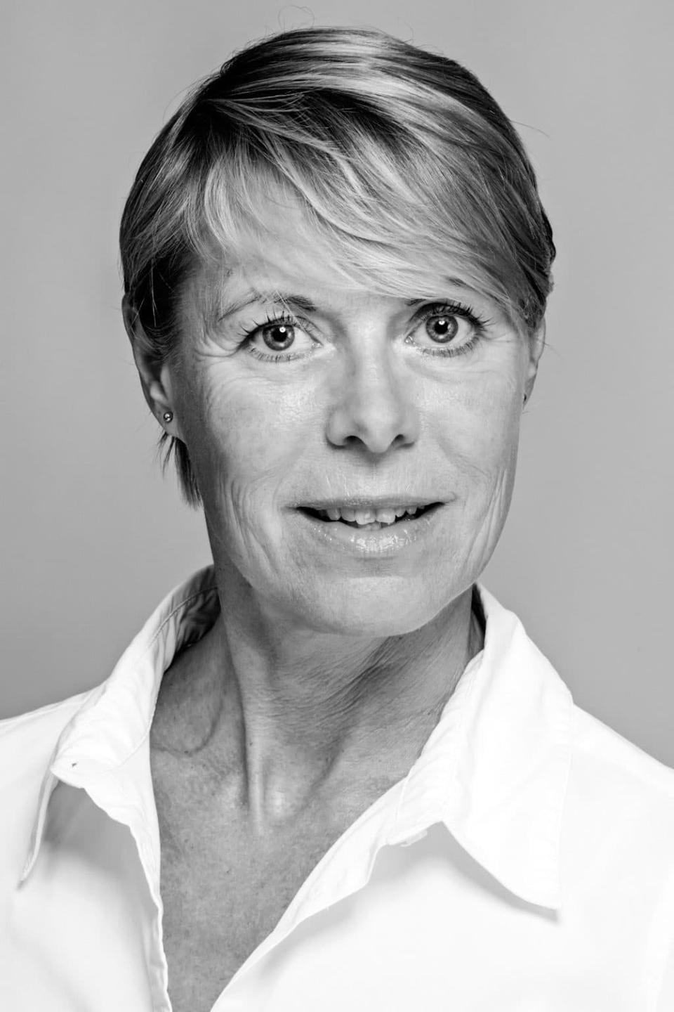 Sort/hvid portrætfoto af kvinde med kort lyst hår og let smil i hvid skjorte mod en lys baggrund. Fokus på ansigtet.
