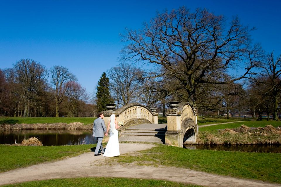 Bryllupsfoto af brudepar hånd i hånd på en grussti foran en stenbro over en sø i slotshaven i forårssolen.