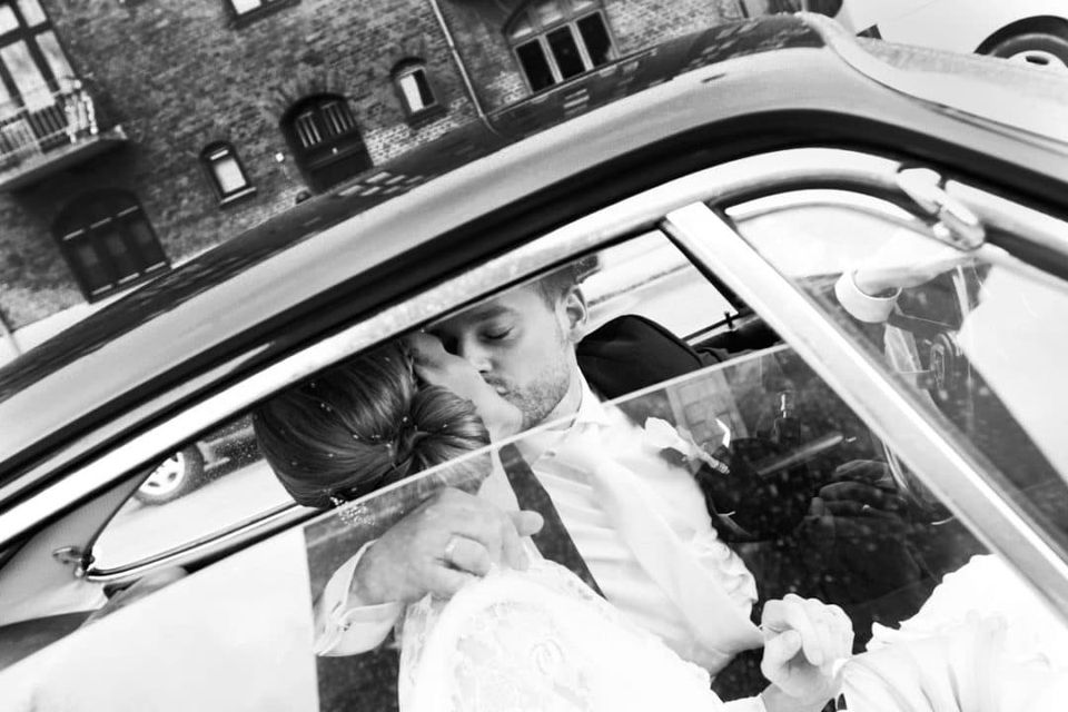 Bryllupsfoto i sorthvid af brudepar i en bil; brudgommen kysser bruden på panden, set gennem bilruden.