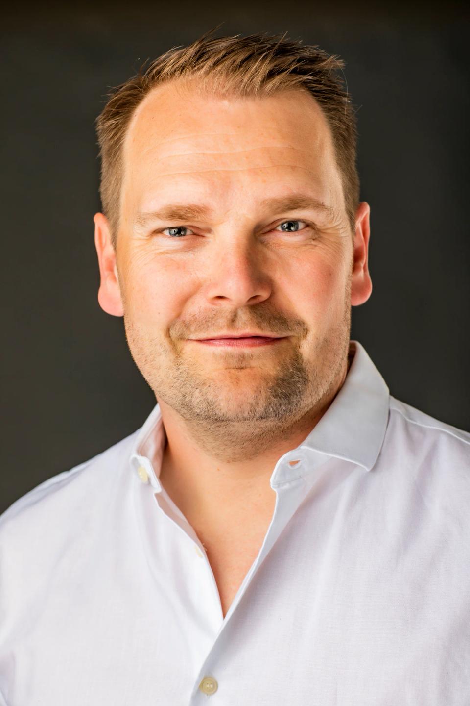 Portrætfoto af smilende mand i hvid skjorte med kort skæg og venlige øjne mod mørk baggrund.