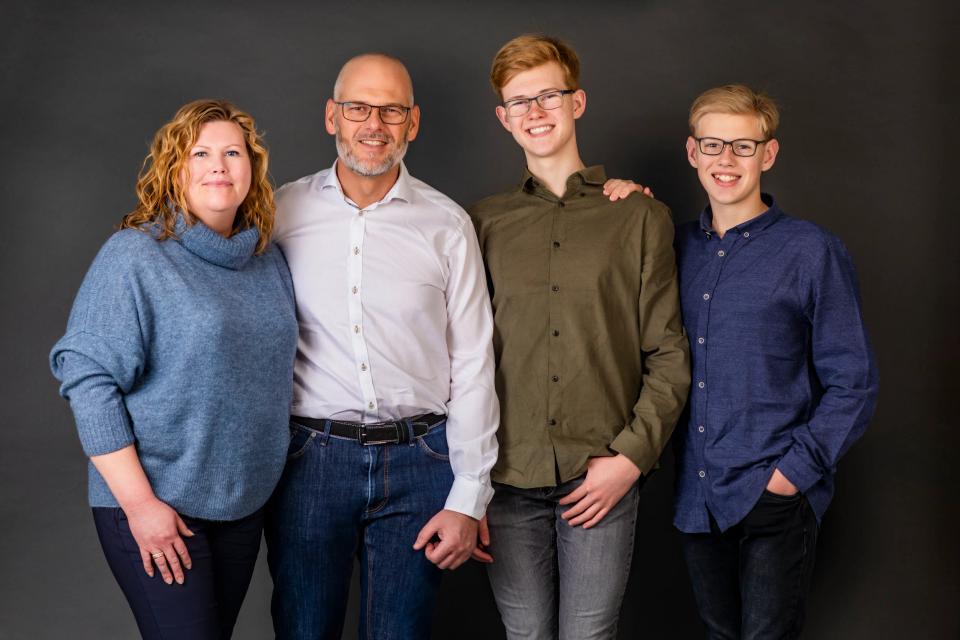 Hjertevarmt familiefoto af mor, far og to glade sønner i Virum, smilende mod kameraet på mørk baggrund.