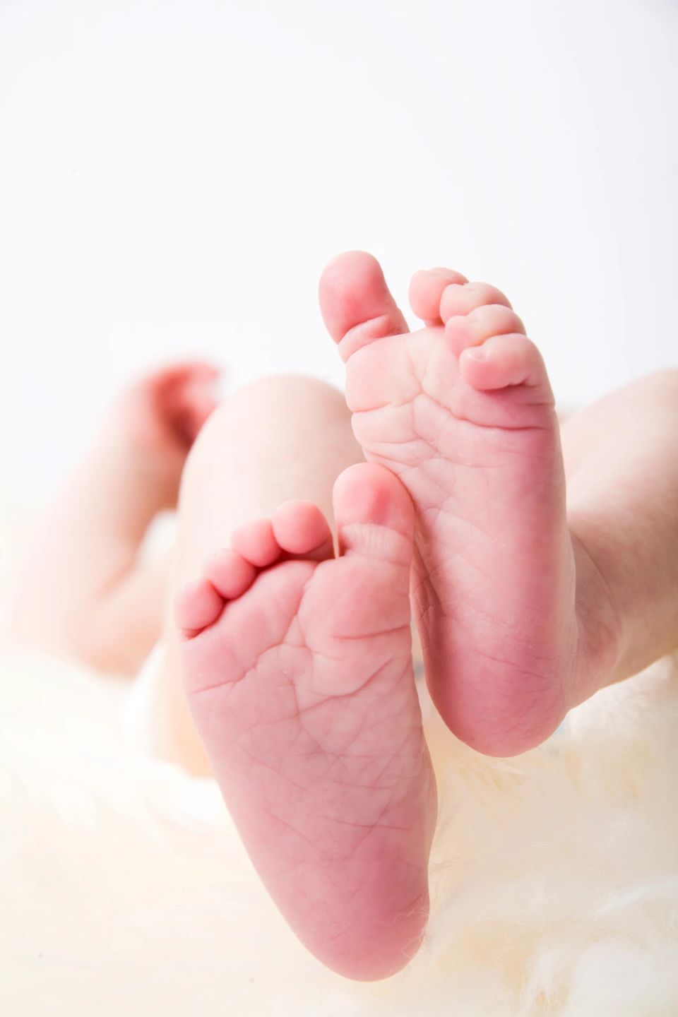 Nært babyfoto af små lyserøde babyfødder med bløde folder liggende på et hvidt fluffy tæppe i naturligt lys.