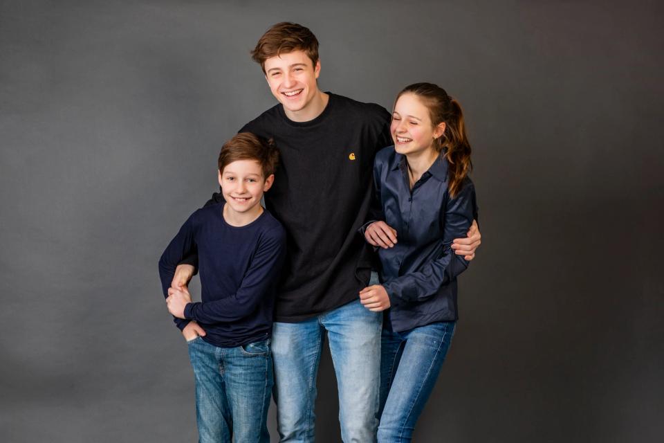 Tre glade søskende i levende familieportræt mod grå baggrund. Midterste dreng smiler bredt, pigen griner, og den lille dre...