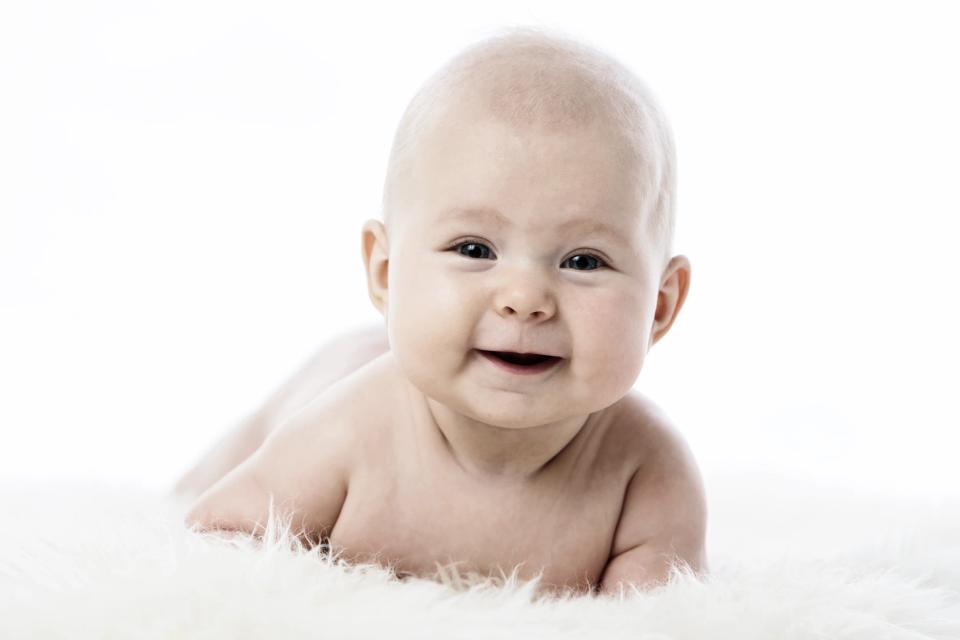 Nærbillede af glad baby med åben mund og store øjne, der ligger på et hvidt, blødt tæppe i et studio. Baby fotografering s...