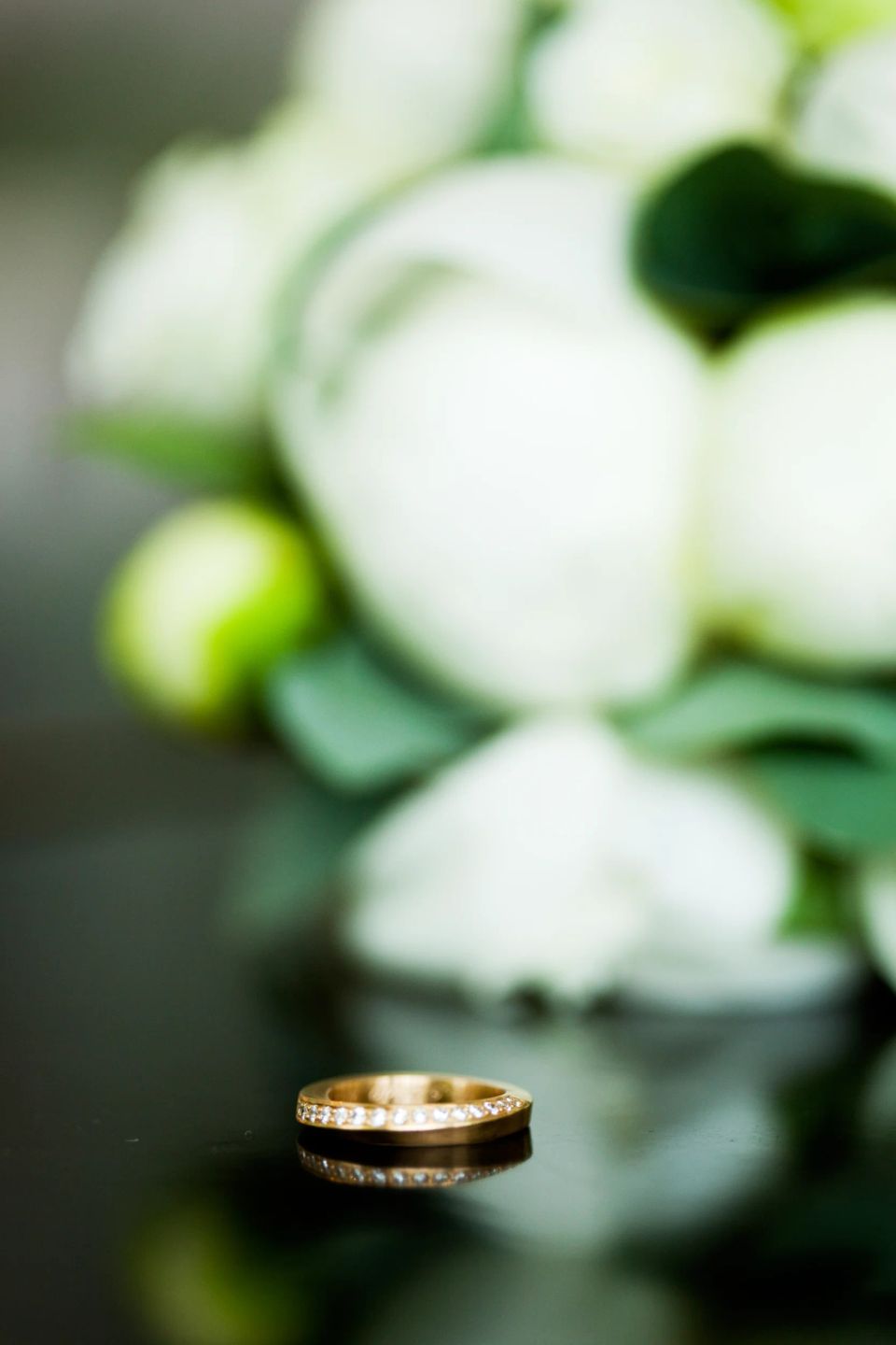 Bryllupsfotografering af en diamantbesat vielsesring i guld, der reflekteres på en mørk overflade med hvide blomster i blø...